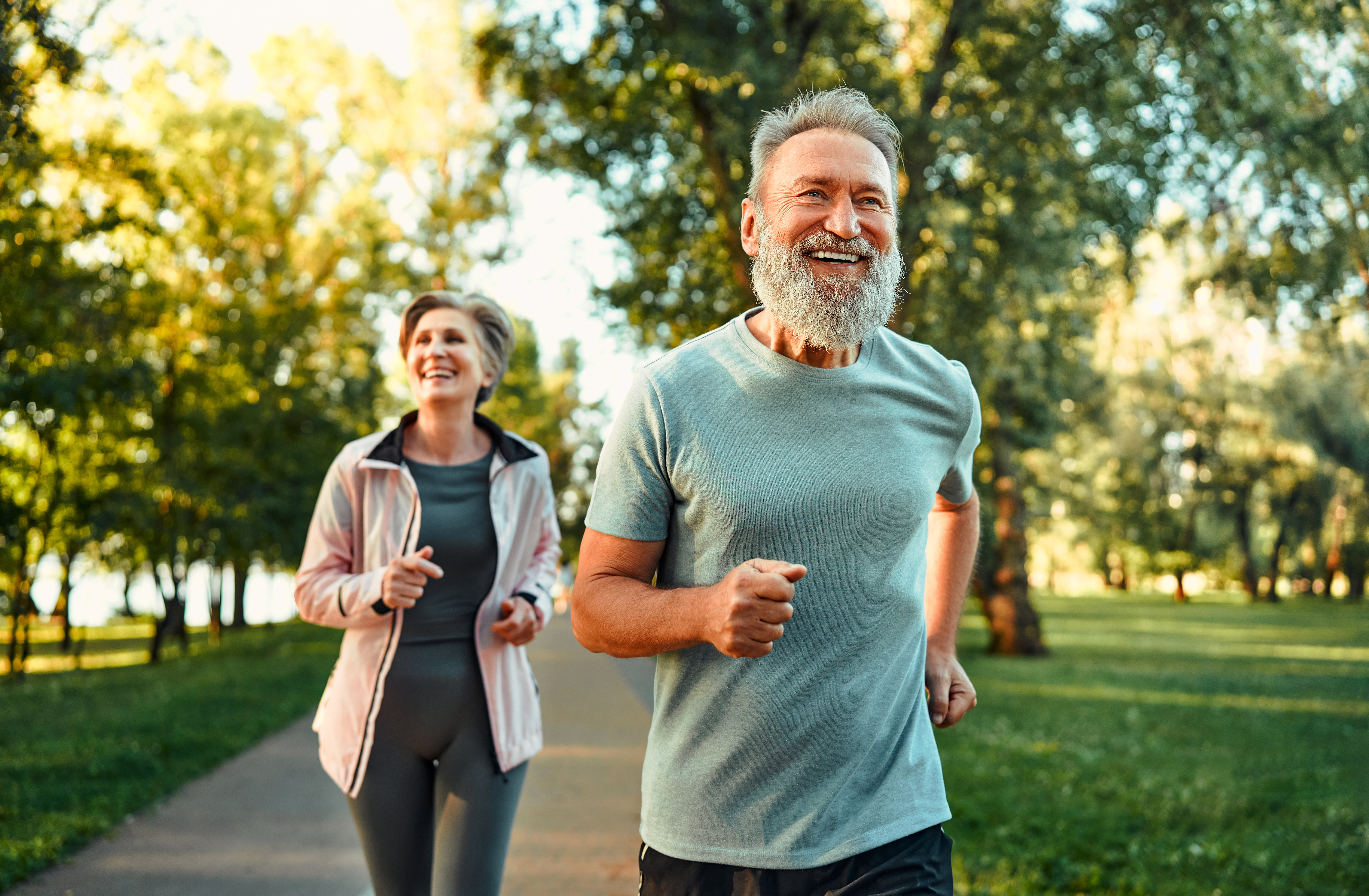 A man and woman jogging happily on a tree-lined path in sunlight. The man wears a blue shirt, the woman a pink jacket. Both are smiling.