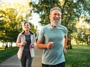A man and woman jogging happily on a tree-lined path in sunlight. The man wears a blue shirt, the woman a pink jacket. Both are smiling.