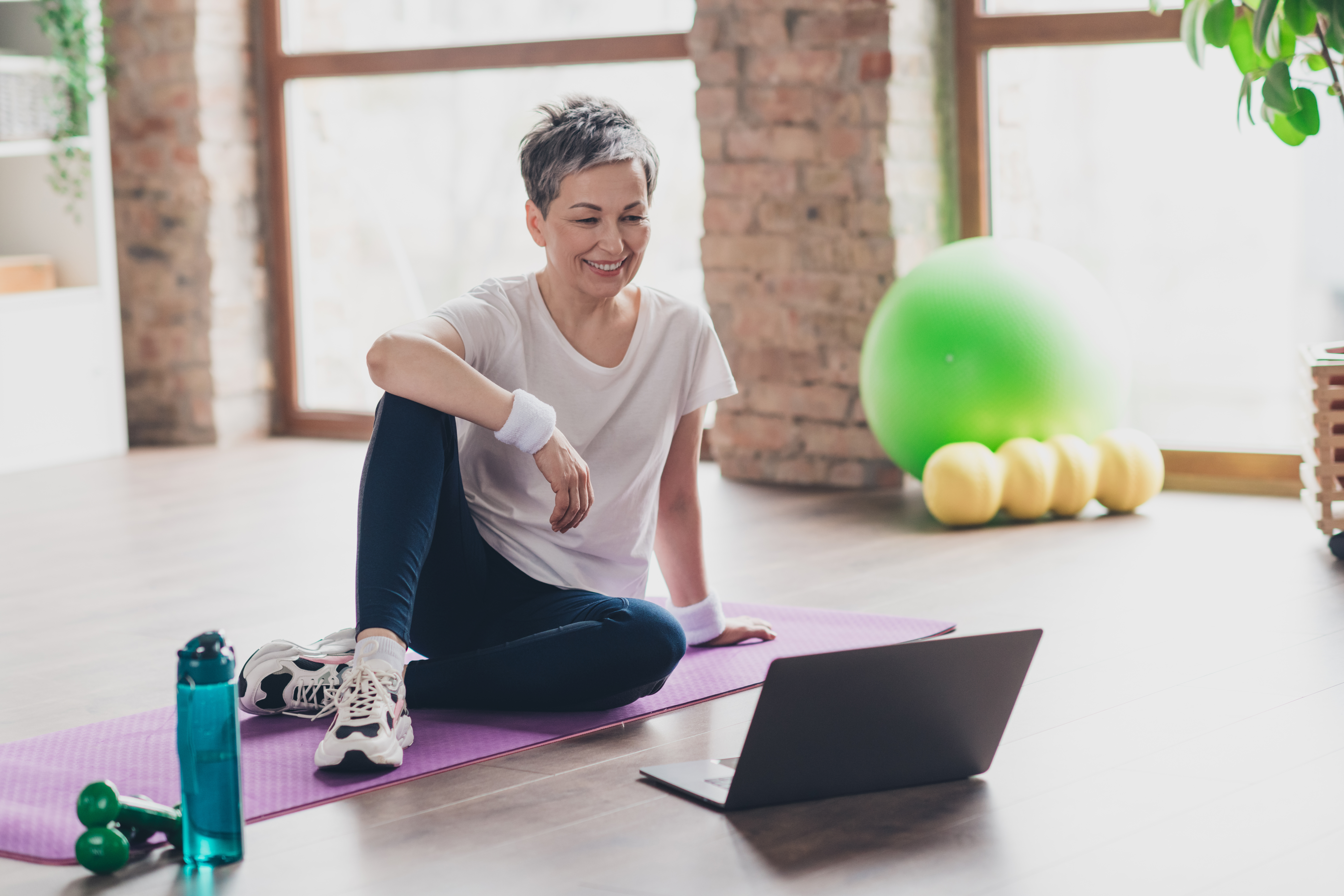 Woman smiling, sitting on a purple mat, watching a laptop. Background: brick wall, large window, green exercise ball, yellow weights.