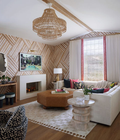 Contemporary living room with a large white sectional sofa and colorful throw pillows, centered around a round tan leather ottoman. The feature wall behind the fireplace is covered in diagonal wood-patterned wallpaper, adding texture and warmth. A round wooden chandelier hangs from a ceiling with exposed wood beams. The room includes a modern electric fireplace, a patterned accent chair, layered rugs, and natural-toned side tables. Tall windows are framed with white curtains edged in pink, letting in ample light.
