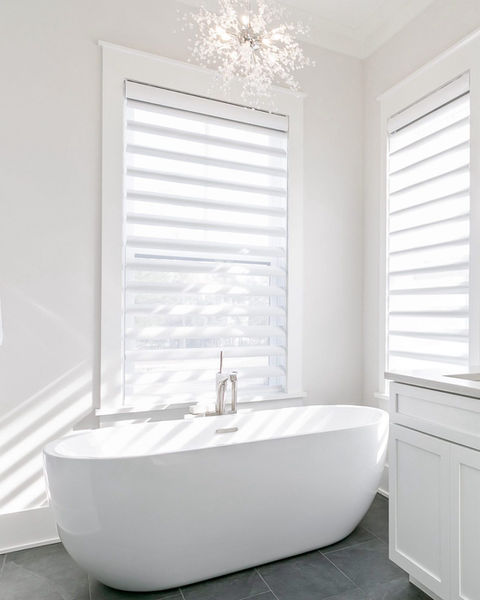  Minimalist bathroom with a modern freestanding white bathtub centered in front of a large window. The space is filled with natural light filtered through white zebra blinds that create soft shadow patterns across the tub and floor. Light gray tiles and crisp white walls enhance the clean, airy atmosphere, while a subtle, elegant chandelier hangs from the ceiling.