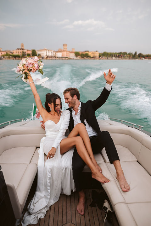 bride and groom ride a boat at sunset with bouquet held high in the air