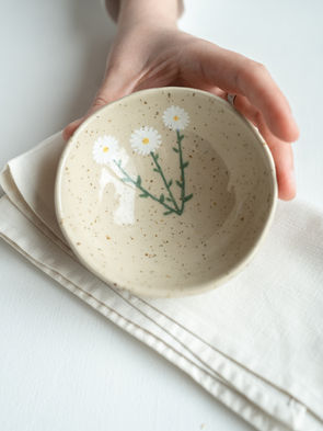 Bildet viser en håndlaget skål i lys leire. Skålen er håndmalt med motiv av prestekrager. 