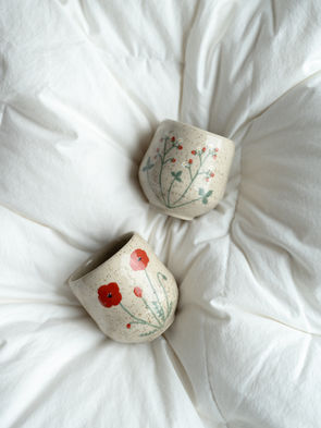 Bildet viser en håndlaget kopp i lys leire. Koppen er håndmalt med motiv av markjordbær og valmuer.