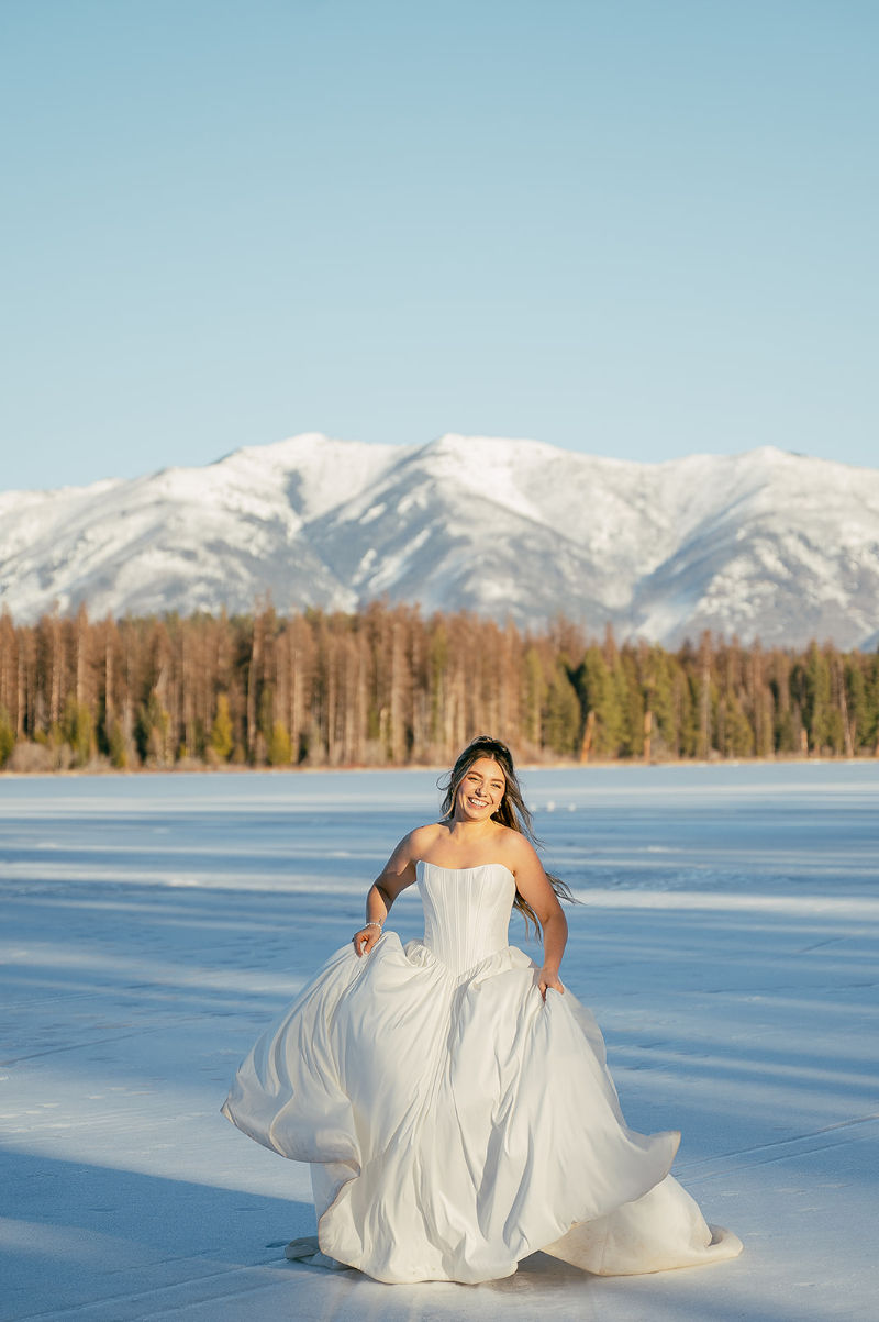 Smiling bride in a flowing white wedding dress running on a frozen lake with snowy mountains, pine trees, and blue sky behind her.