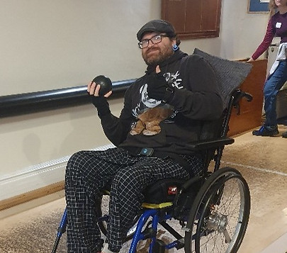 A person in a wheelchair holds a bowling ball, preparing to bowl, with a bowling alley visible in the background.