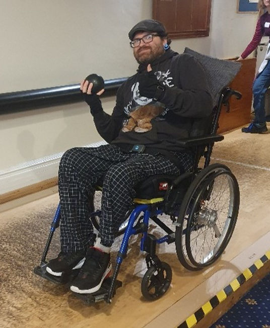 A person in a wheelchair holds a bowling ball, preparing to bowl, with a bowling alley visible in the background.