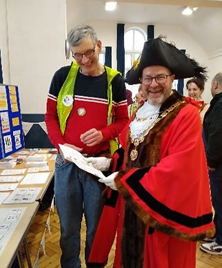 A person in traditional mayoral regalia stands beside a casually dressed individual, reviewing documents at a community event.