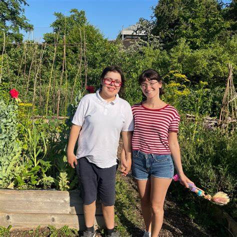 A girl and a woman stand in a garden. There are lots of plants and trees behind them. The woman is holding a doll.