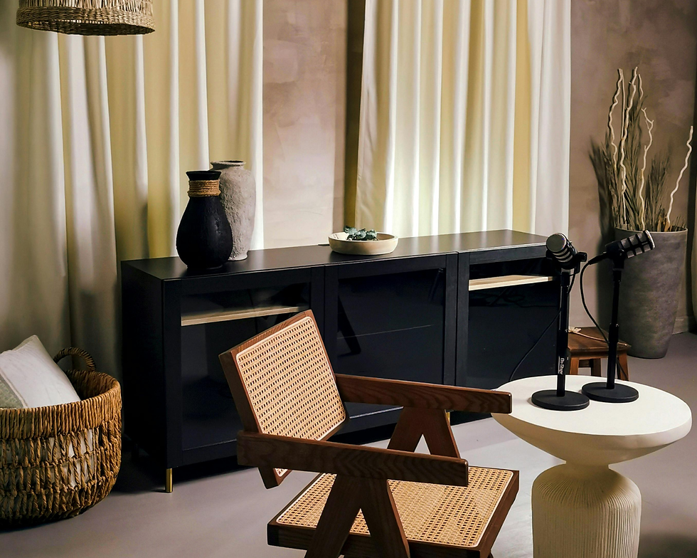 Estúdio aconchegante com cadeira de palha, mesa branca e dois microfones, diante de armário preto e cortinas claras.