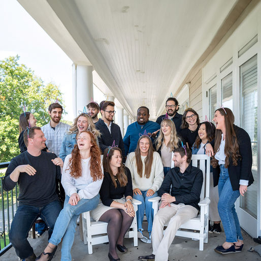 A group of people wearing unicorn headbands laugh together on a porch. They're dressed casually, creating a joyful and lively atmosphere.