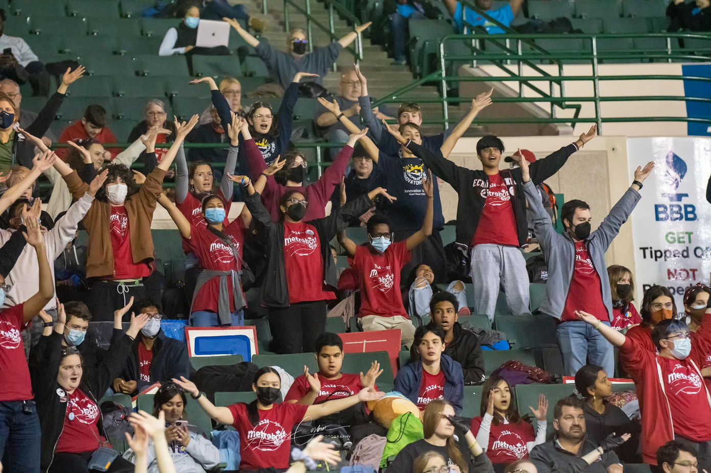 Excited fans at a robotics competition cheer with arms raised high.