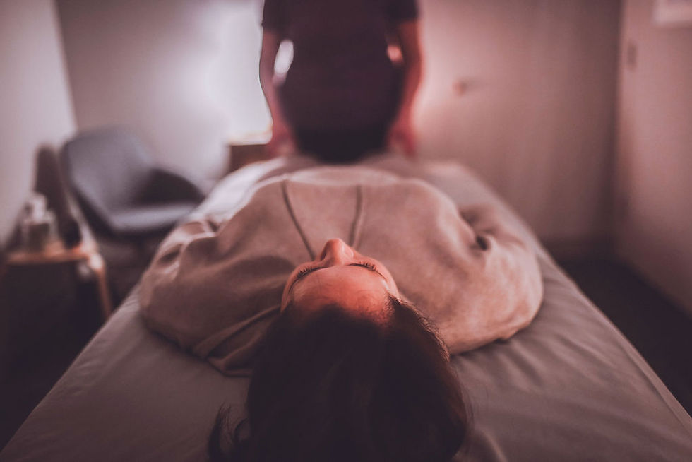 Person lying on a massage table, another person stands nearby. Warm lighting, cozy room with a chair. Relaxing atmosphere. craniosacral therapy session at Nourish and Align Baltimore