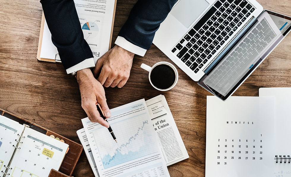 Hands in a suit reviewing financial charts and graphs on a wooden desk with a laptop, coffee, newspaper, and calendar. Professional setting.