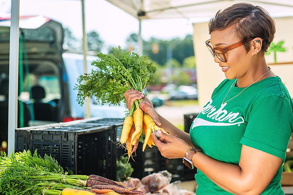 Ja'Nell in a green shirt that says Durham. She is holding colorful locally grown carrots at a local farmers' market.