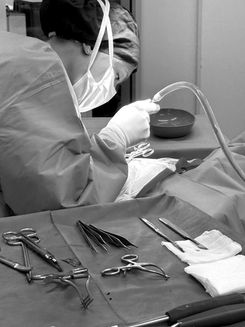 OP-Tisch mit chirurgischen Instrumenten, daneben ein Operateur mit Gesichtsmaske und dunkler Kopfbedeckung beim Absaugen von einer Flüßigkeit.