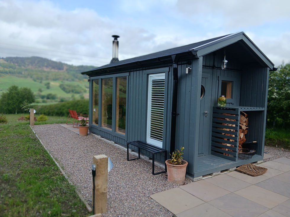 Modern gray sauna cabin with wood stove and stacked firewood on a gravel path, set in green hills under a cloudy sky