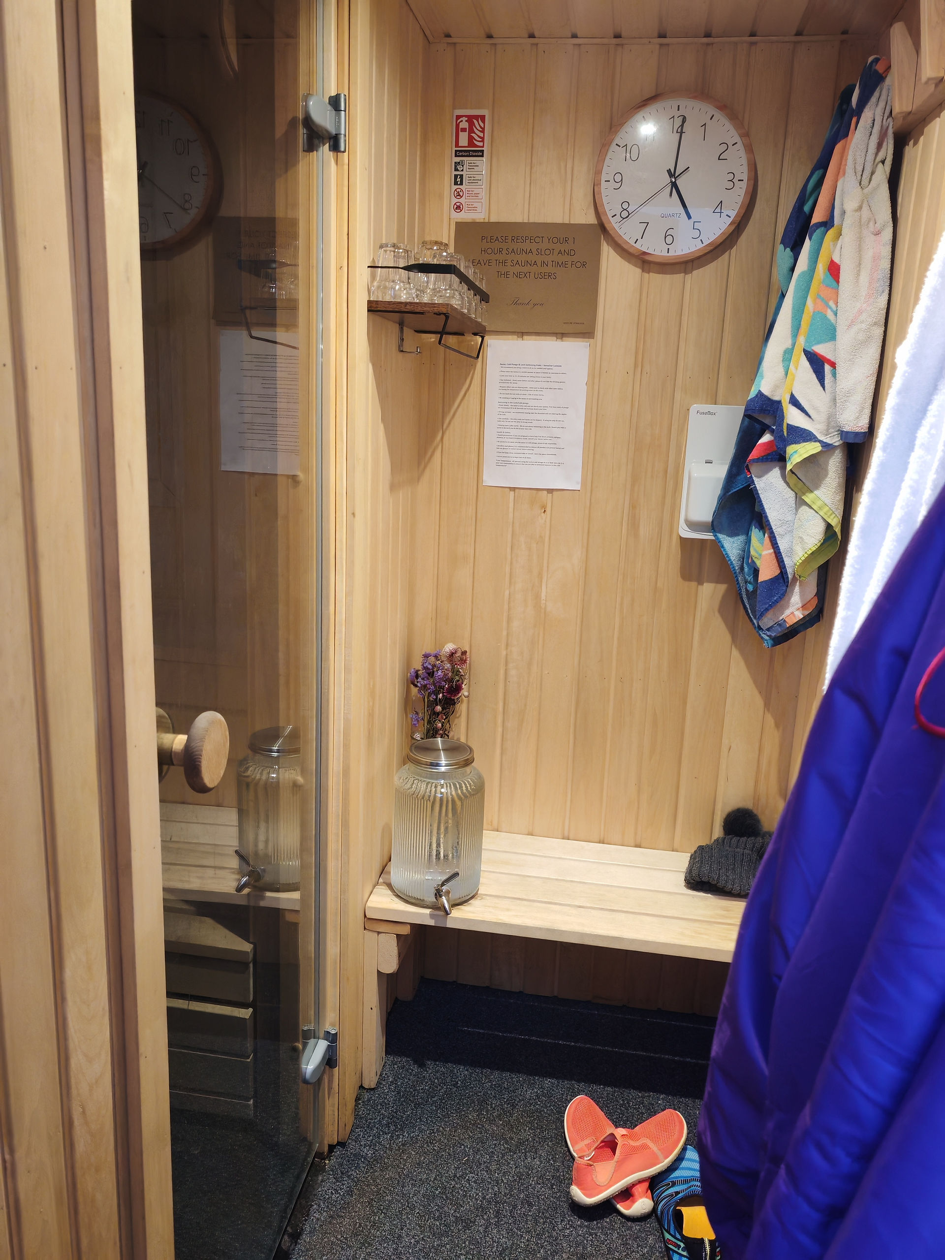 wooden entry area to sauna with towels and jackets hung up from hooks with clock on wall
