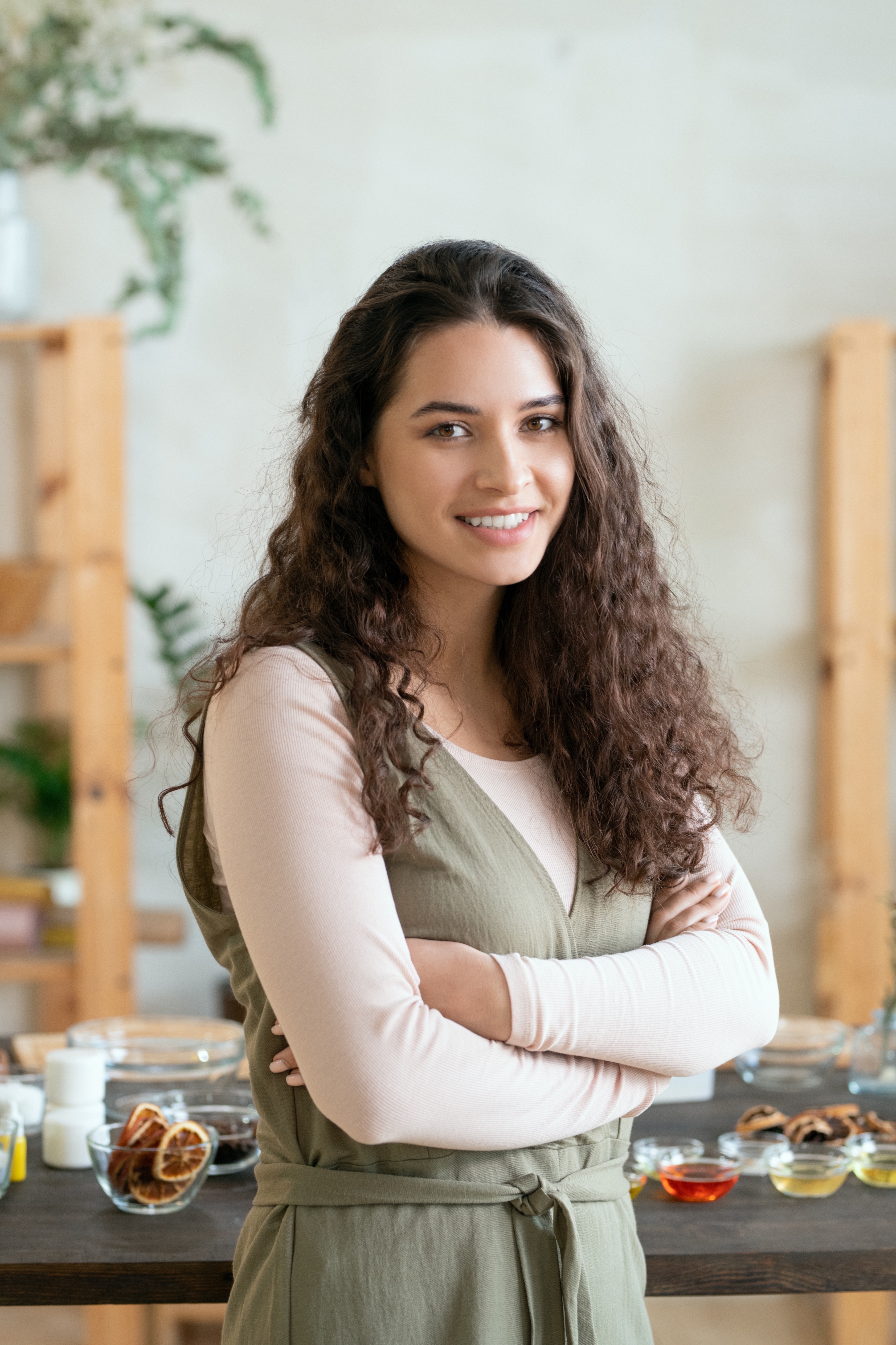 young-smiling-brunette-woman-crossing-arms-by-chest-while-standing-against-table-with-ingr