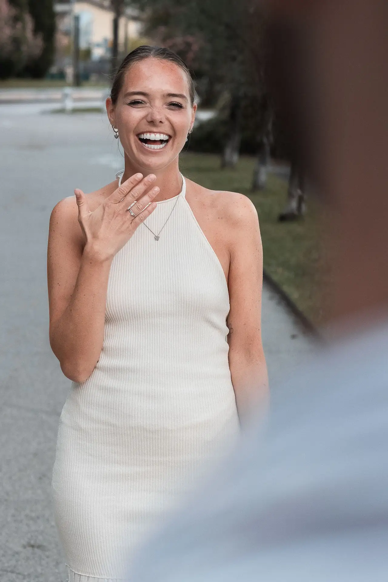 Young woman smiling while admiring her hands, showing off engagement rings, wearing a white halter dress.