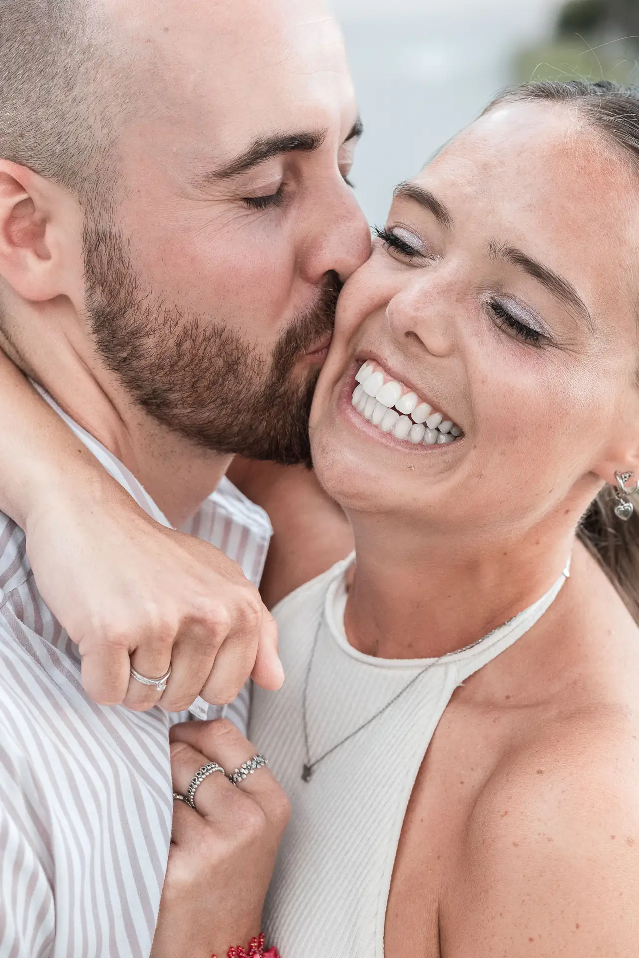 Happy couple embracing, the man kissing the woman on the cheek while she smiles joyfully showing her ring.