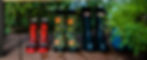 Four pairs of rain boots sit on a wooden deck. From left: red ladybug, floral, black with "Hunter" label, blue striped. Green plants in background.