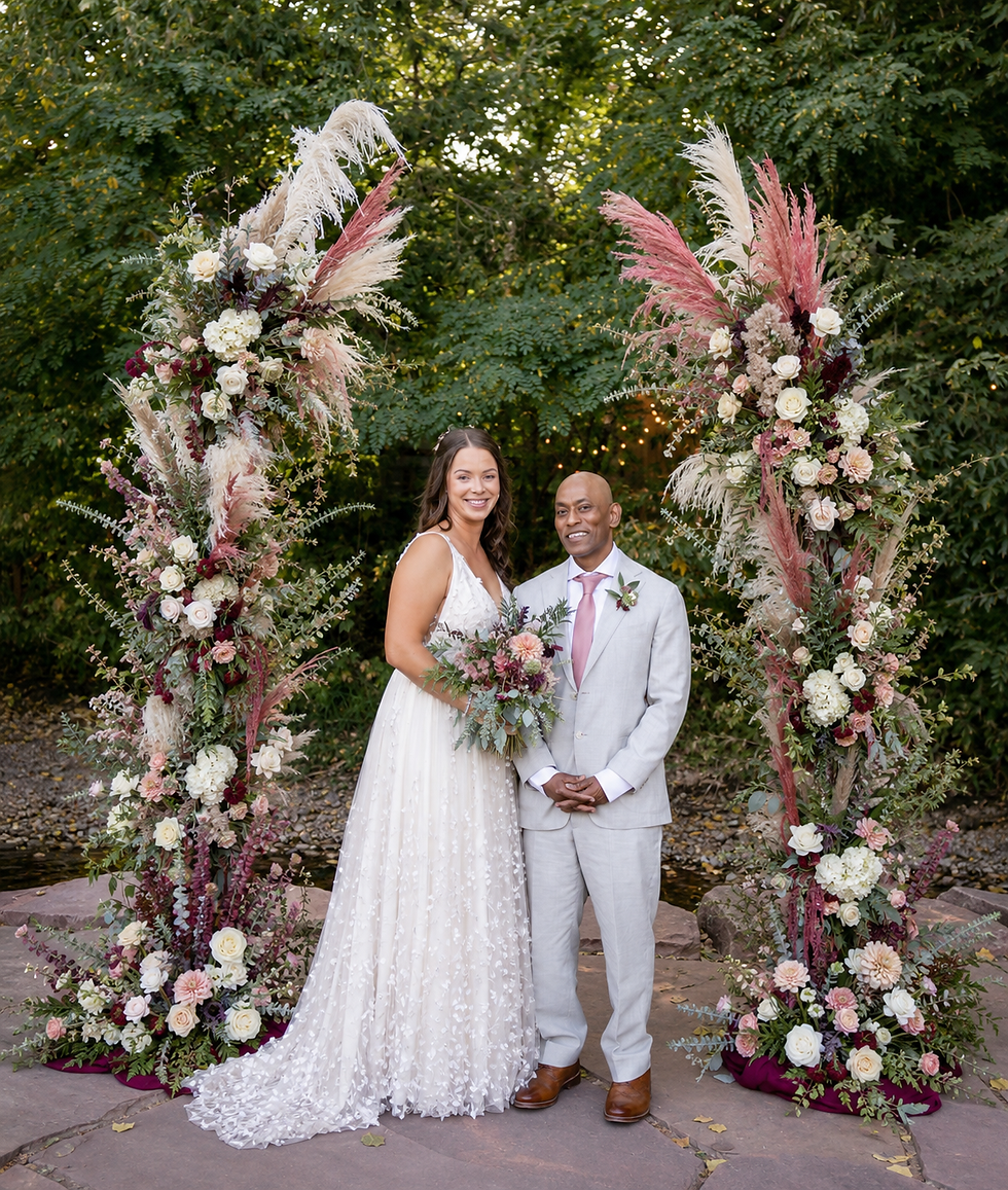 Bride in white gown and groom in light suit pose under pastel floral arch outdoors, smiling, with trees behind. stunning floral arch by petals boulder at the lyons farmette
