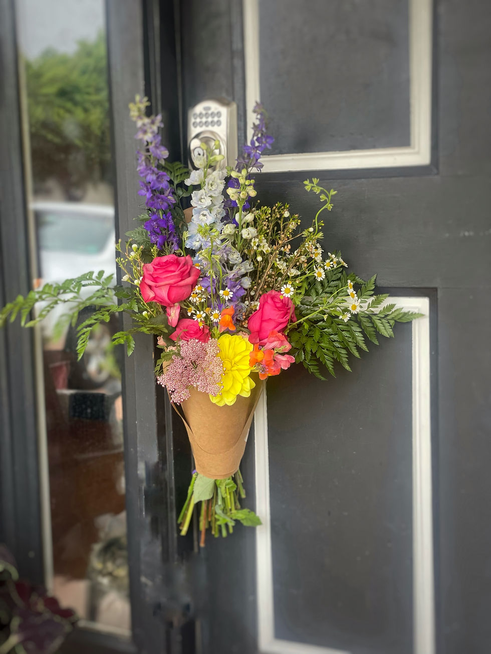 Bright flower bouquet with pink roses and yellow blooms hangs on a black door. A keypad lock is visible. Natural, cheerful setting.