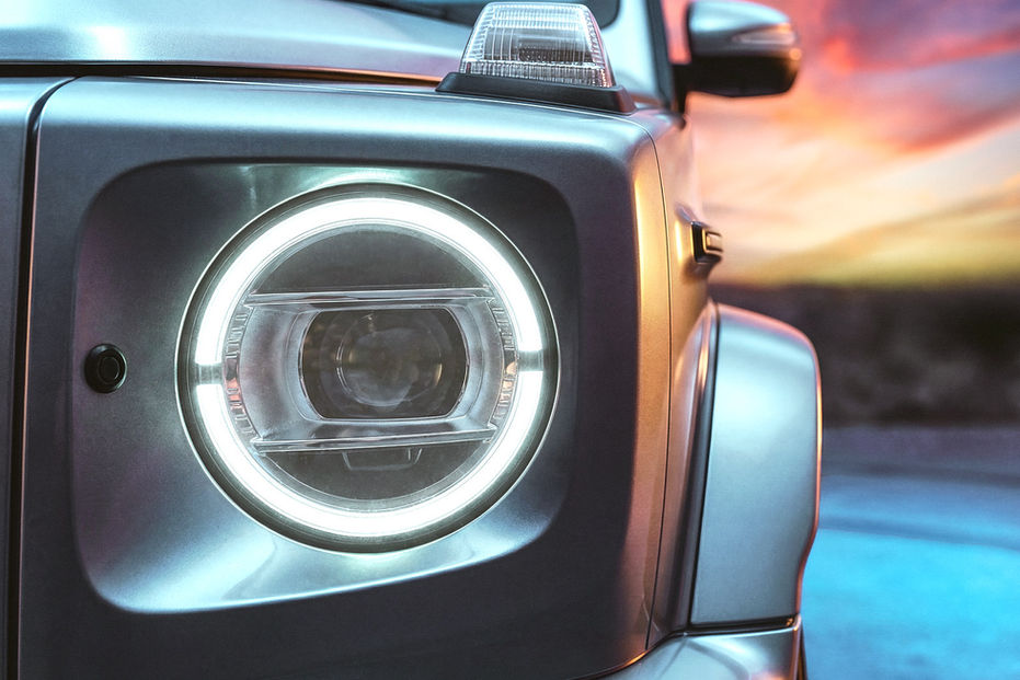 Close-up of the front headlight and bodywork of a silver Mercedes-Benz G-Class luxury SUV at sunset.