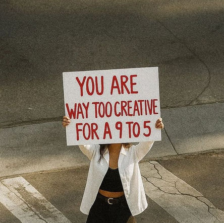 A person standing on a crosswalk holding a sign that reads “You are way too creative for a 9 to 5,” wearing a white shirt, black outfit, and boots, with their face hidden behind the sign.