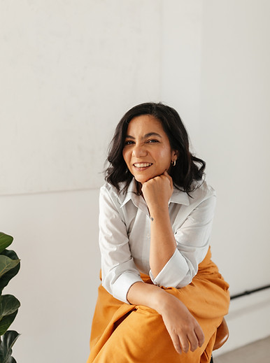 Mulher sentada com mão no queixo sorrindo. É uma foto da psicóloga