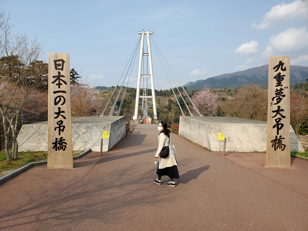 女性が日本一の大吊橋を歩く。晴れた空、周囲に桜、背景に山が見える。橋の入り口には大きな文字の看板が立つ。
