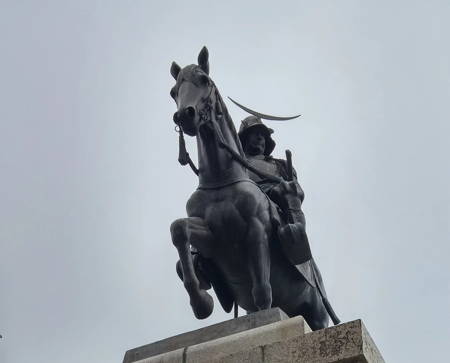 馬に乗った武士の銅像が曇り空を背景にそびえ立つ。武士は兜をかぶり、威厳を感じさせる表情をしている。