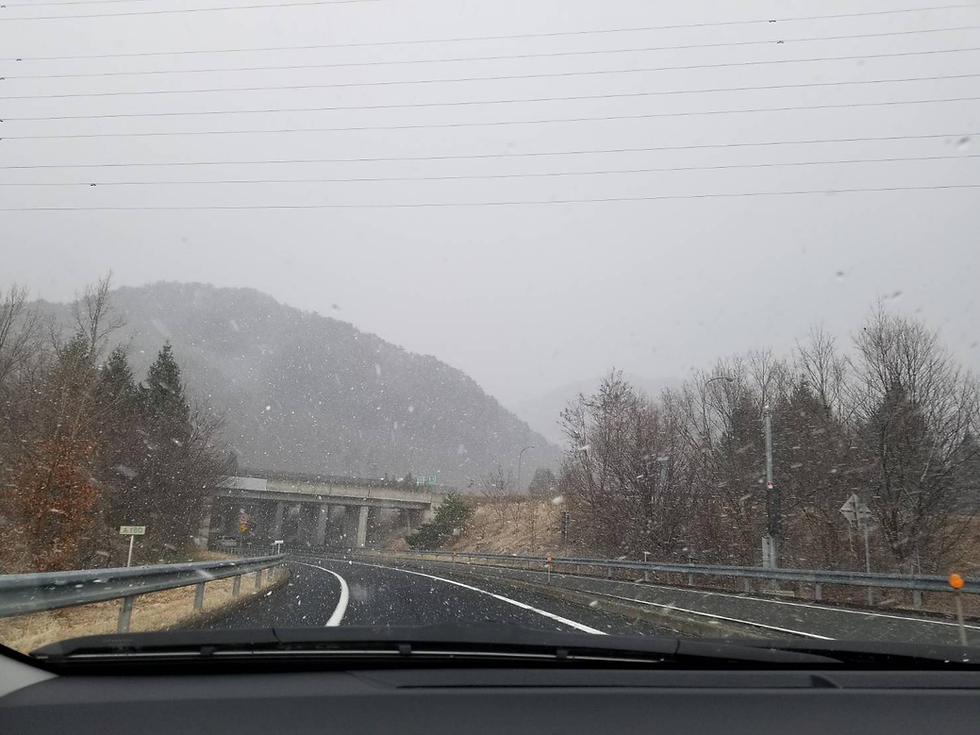 雪が降る山道を車が走行中。道路は曲がりくねり、背景には雪に覆われた山々が見える。曇った空の下、冬らしい景色。