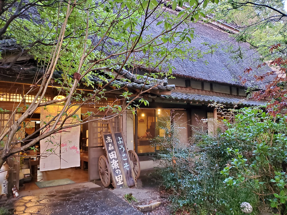 木々に囲まれた茅葺き屋根の和風建物。暖かな灯りがともり、入口に高森田楽の里とようこその看板が立つ。