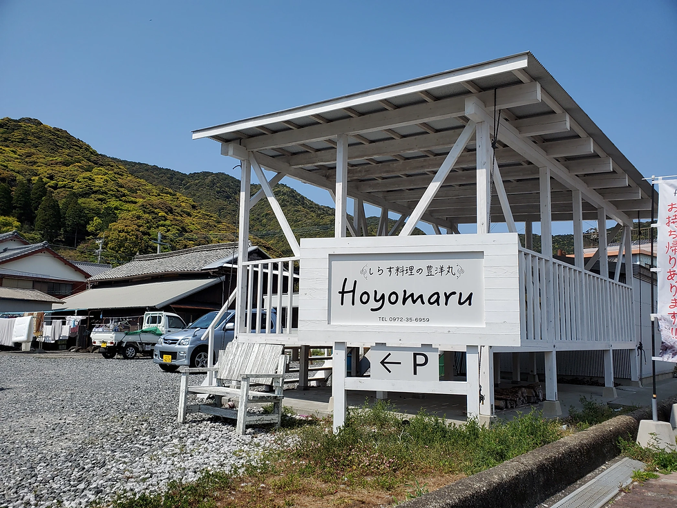 青空の下、山を背景にした白い木造の施設。看板にHoyomaru、下に左矢印とP、静かな港町の雰囲気。