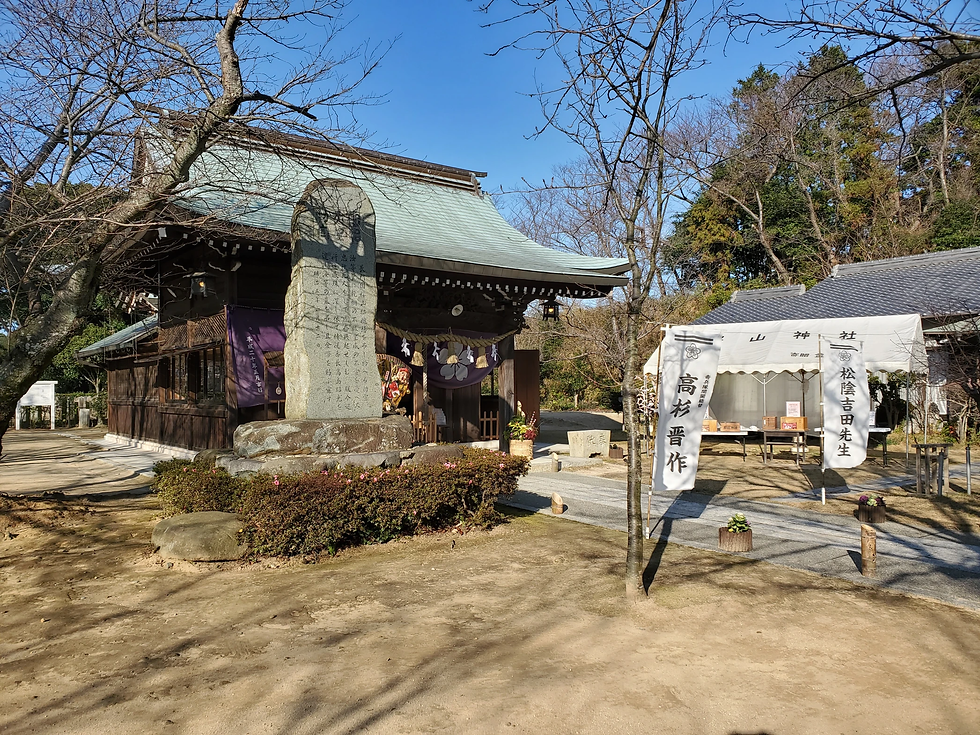 木造の神社、石碑と白い幕に文字がある。参道沿いに緑が広がり、背景には木々が見える。晴天で穏やかな雰囲気。
