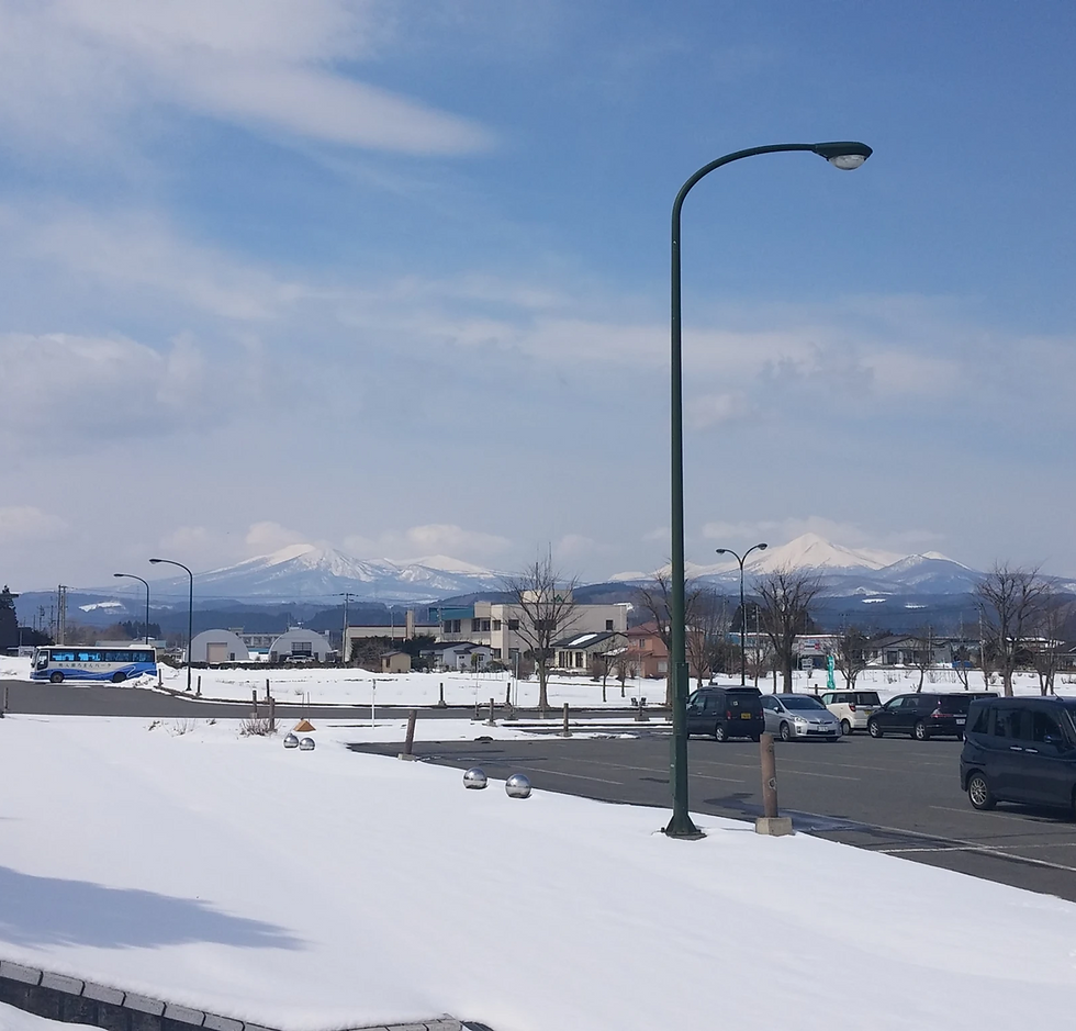 雪の駐車場の背景に山が見える。青空の下、街灯と駐車中の車が並び、遠くに青いバスが走っている。静かな冬景色。