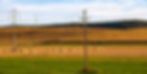 Power lines cross a rural landscape with hay bales on golden fields. A green foreground and wooded hills in the background under a blue sky.