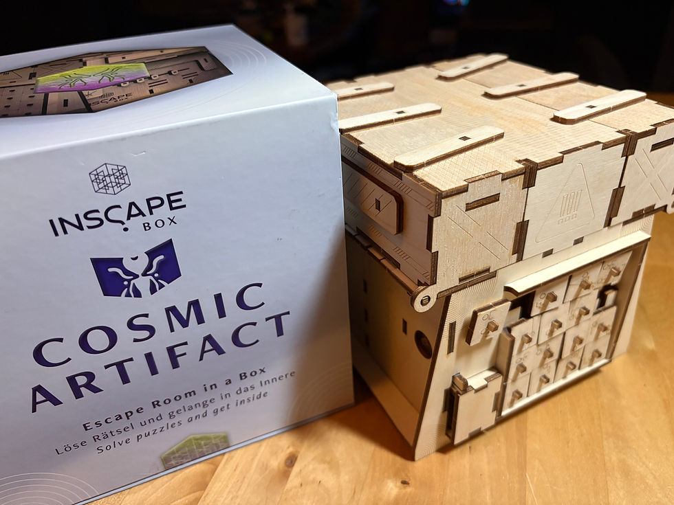Cosmic Artifact by INSCAPE Box on a wooden surface. On the left, the white product box displays the Inscape Box logo, a purple stylized figure emblem, and the text 'Cosmic Artifact — Escape Room in a Box' in English and German. On the right, the assembled wooden puzzle box sits beside it, showing laser-cut plywood panels with engraved symbols, sliding mechanisms, a grid of small push tabs, and a partially visible latch along one edge.