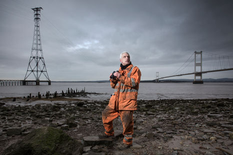 Portrait of linesmen with binoculars standing on a rocky shore, inspecting the electricity cables that cross high above the Severn Estuary. 