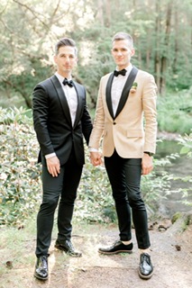 A cozy, intimate moment between two grooms dressed in deep burgundy suits, sitting together in a warm, vintage-style venue.