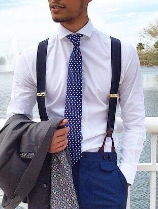 A groom in a white dress shirt with navy suspenders and a polka dot tie, holding his suit jacket over his shoulder while standing in front of a fountain, blending formal and casual wedding fashion.