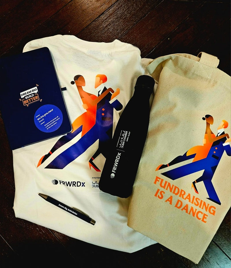 FRWRDx IDEA Program graduation kit laid flat — branded t-shirt, tote bag reading "Fundraising is a Dance," notebook, water bottle, and pen on a dark wooden surface