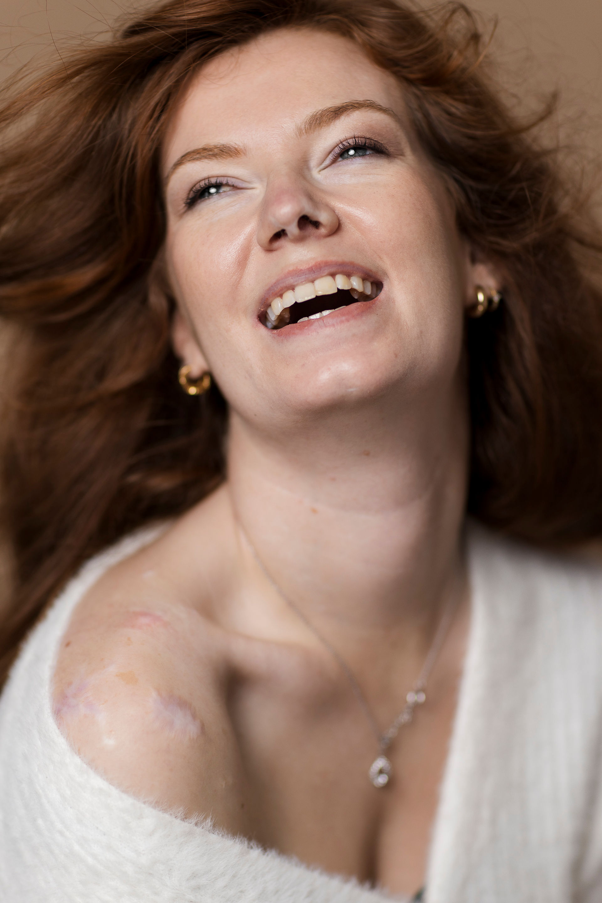 Femme rousse sur fond beige avec un pull écru qui rigole