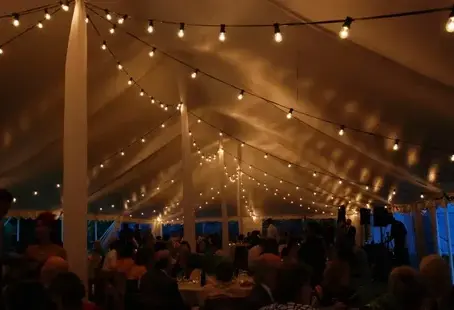 Outdoor event under a large tent decorated with warm string lights and guests seated at tables in the evening