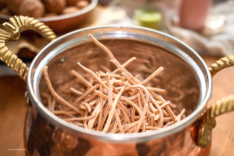 Copper bowl with brass handles holding thin dried strips of pasta on a wooden table; © serra moon food watermark.