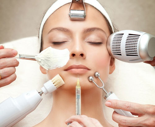 Woman receiving facial treatments with various beauty tools in a spa setting. Calm expression, white headband, light background.