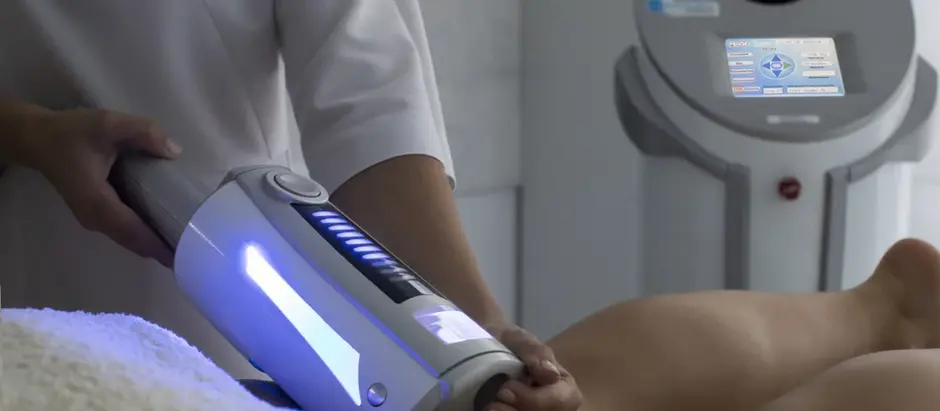 Person using a gray cellulite treatment machine on a patient's leg in a clinical setting, with a glowing blue display. Calm atmosphere.