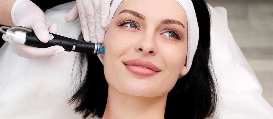 Woman with dark hair receives a facial treatment with a device, smiling slightly. Wears a white headband, gloved hands visible. Relaxed setting.